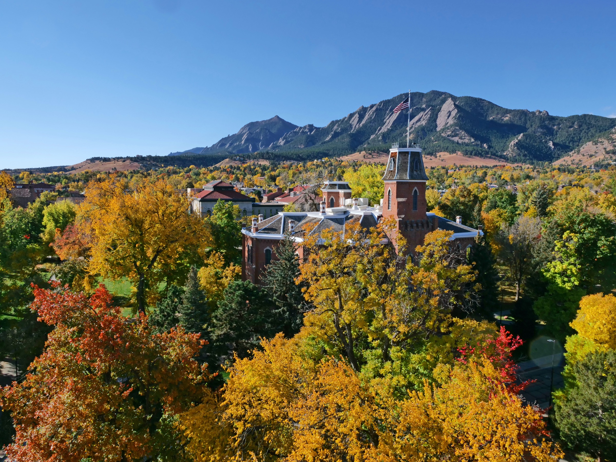 Autumn trees surrounding a historic CU Boulder campus building with the Flatirons behind it.