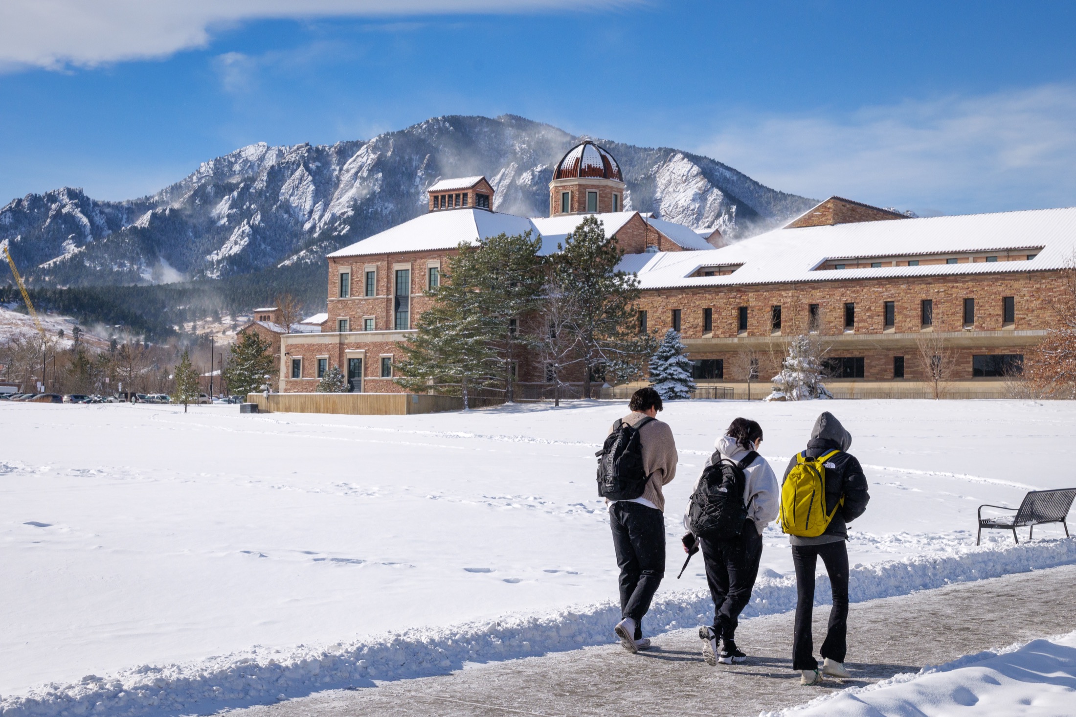 Students walking past a snow-covered CU Boulder building with mountains behind it.